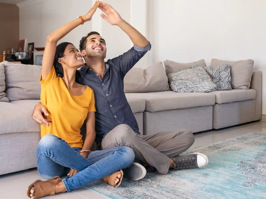 a couple sitting next to each other next to couch holding their hands together to make a house roof above them